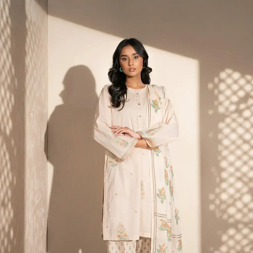 Woman wearing a light-colored embroidered outfit standing against a textured wall.